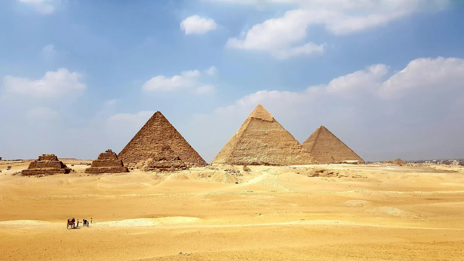 Die Pyramiden von Gizeh in der Wüste unter blauem Himmel, im Vordergrund winzige Figuren und ein Pferdewagen als Größenvergleich
