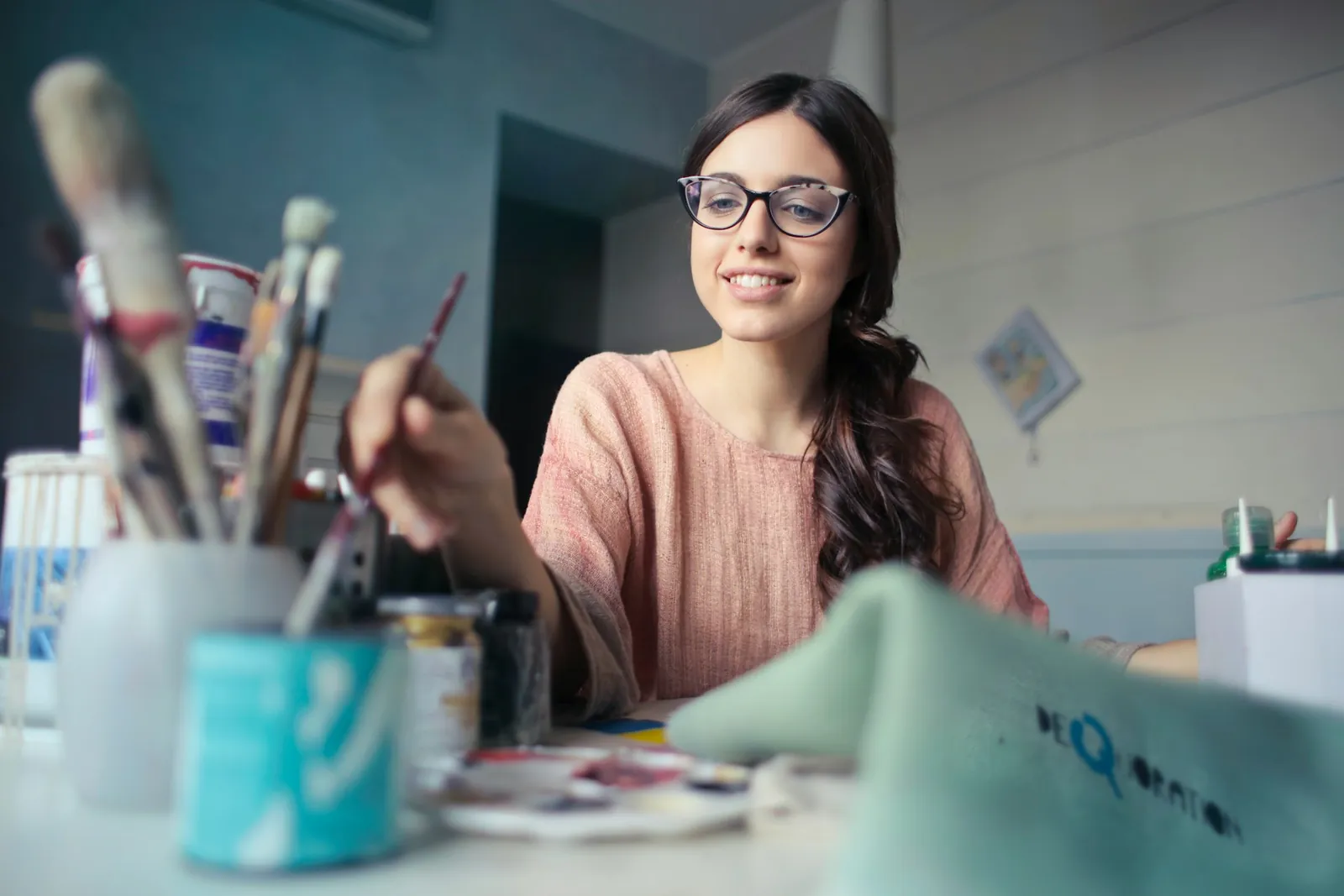 Eine junge Frau mit Brille lächelt, während sie an einem Tisch voller Pinsel und Farbtöpfe malt, konzentriert und ihrem Handwerk gewachsen